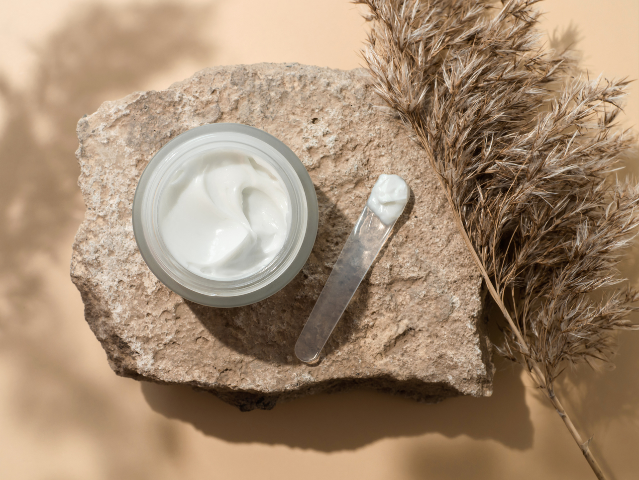 Tarro de crema facial con espátula sobre piedra decorativa y fondo beige